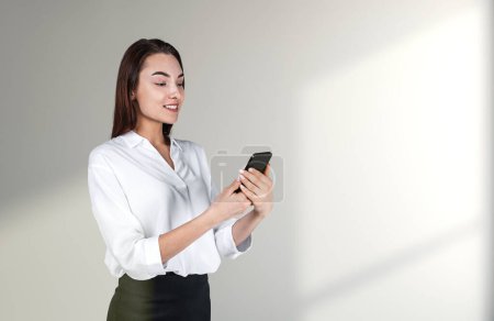 Foto de Young attractive businesswoman wearing formal wear is standing holding smartphone near concrete wall with sun light in background. Concept of working process on mobile gadget, internet communication - Imagen libre de derechos