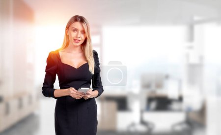 Foto de Young attractive businesswoman wearing formal wear is standing holding smartphone at office workplace with sun light in background. Concept of working process at workspace, internet communication - Imagen libre de derechos