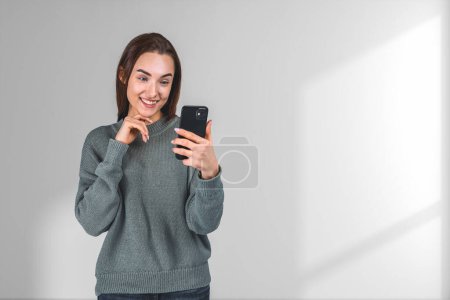 Téléchargez les photos : Smiling attractive businesswoman in casual wear standing holding smartphone near empty white wall in background. Concept of inspired business person, dreaming woman, social media, mobile application - en image libre de droit