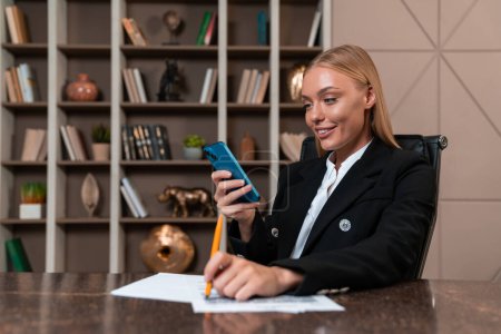 Photo for Smiling businesswoman looking at the with phone, office room with shelf and decor. Business desk with papers and pen. Concept of business network and social media - Royalty Free Image