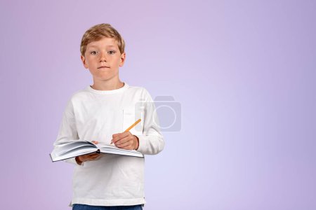 Foto de Pondering handsome boy in casual wear standing holding notebook and taking notes near empty purple wall in background. Concept of inspired kid, education, learning, studying, student - Imagen libre de derechos