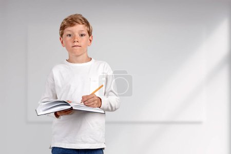 Téléchargez les photos : Pondering handsome boy in casual wear standing holding notebook and taking notes near empty white wall with mockup in background. Concept of inspired kid, education, learning, studying, student - en image libre de droit