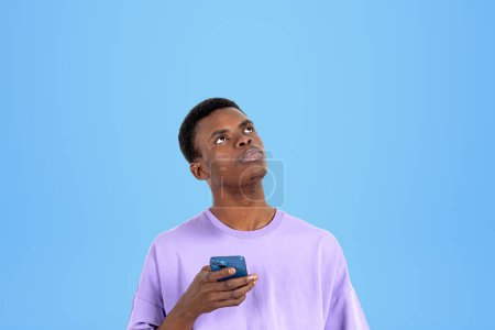 Téléchargez les photos : African man in purple t-shirt with phone in hand, thoughtful look up on empty copy space blue background. Concept of mobile app and planning - en image libre de droit