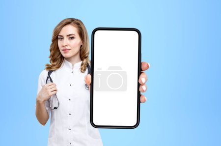 Photo for Young doctor woman show smartphone, blue background. Concept of telemedicine and consultation. Mock up copy space display - Royalty Free Image