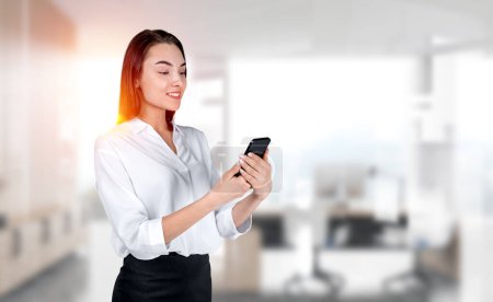 Photo for Young attractive businesswoman wearing formal wear is standing holding smartphone at office workplace with sun light in background. Concept of working process on mobile gadget, internet communication - Royalty Free Image