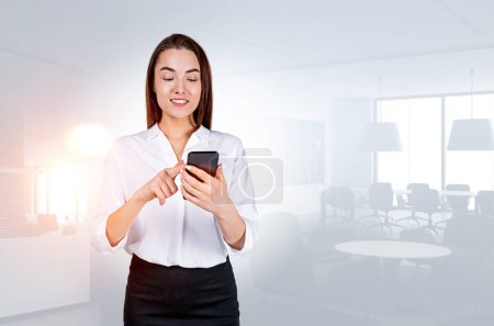 Photo for Young attractive businesswoman wearing formal wear is standing holding smartphone at office workplace with armchairs in background. Concept of working process on mobile gadget, internet communication - Royalty Free Image