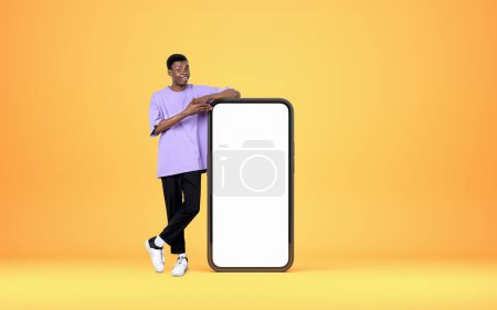 Téléchargez les photos : Young black man smiling, full length, pointing at smartphone mock up copy space screen, yellow background. Concept of online network and social media - en image libre de droit