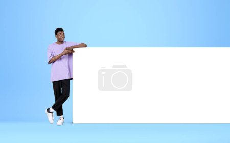 Photo for African American young man finger pointing at mockup huge poster on light blue background. Blank white canvas. Concept of presentation and ad - Royalty Free Image