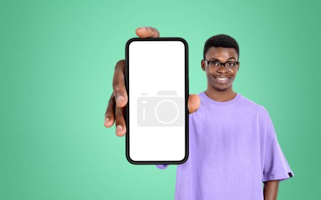 Téléchargez les photos : Smiling black man in eyeglasses holding phone with mock up copy space screen, light green background. Concept of network and social media - en image libre de droit