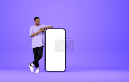 Téléchargez les photos : Young african man smiling, pointing at smartphone mock up copy space screen, purple background. Concept of new website and mobile app - en image libre de droit