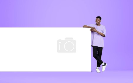 Foto de Joven afroamericano señalando con el dedo el enorme póster de maqueta sobre fondo violeta claro. Lienzo blanco en blanco. Concepto de presentación y anuncio - Imagen libre de derechos