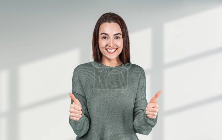 Foto de Joven mujer feliz mostrando el pulgar hacia arriba sobre fondo gris, mirando a la cámara riendo. Concepto de aprobación y similares - Imagen libre de derechos