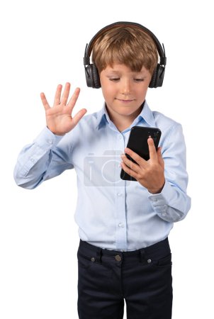 Téléchargez les photos : Smiling child boy in headphones, waving hand and looking at smartphone, isolated over white background. Concept of video conference call and online education - en image libre de droit