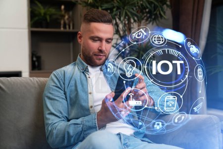 Téléchargez les photos : Man sitting on sofa with smartphone in hands, IOT hud with digital cloud icons and smart devices connection. Concept of artificial intelligence - en image libre de droit