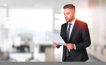 Photo for Portrait of bearded young businessman in elegant suit reading documents in blurry office. Concept of paperwork and leadership - Royalty Free Image