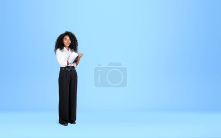 Téléchargez les photos : Femme d'affaires africaine écrivant dans un cahier d'affaires, regardant la caméra et souriant, pleine longueur sur fond bleu clair. Concept d'éducation et de démarrage. Espace de copie. - en image libre de droit