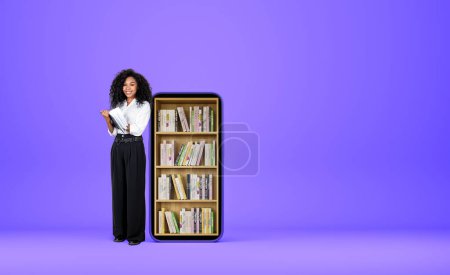 Foto de Mujer de negocios africana sonriendo cerca de teléfono inteligente y tomar nota en el cuaderno. Biblioteca digital, libros en estante sobre fondo púrpura. Concepto de educación en línea. Copiar espacio - Imagen libre de derechos