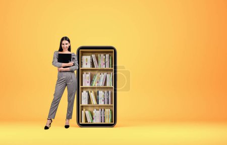 Photo for Smiling businesswoman with notebook standing near smartphone, full length. Digital library, books on rack on yellow background. Concept of online education and courses. Copy space - Royalty Free Image
