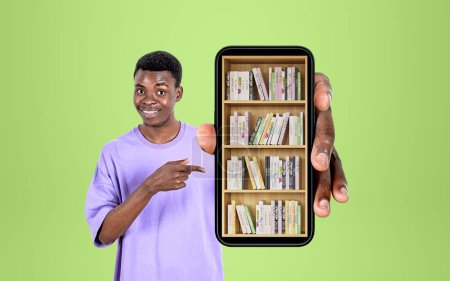 Foto de Hombre africano sonriendo, sosteniendo el teléfono inteligente y el dedo apuntando a la biblioteca digital, libros en el estante, fondo verde. Concepto de educación en línea y aplicación móvil - Imagen libre de derechos