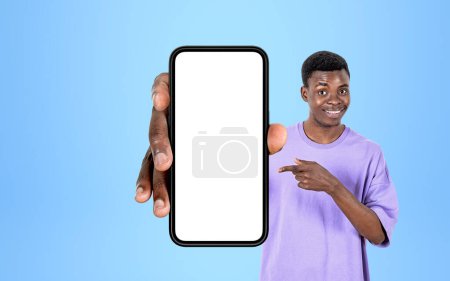 Photo for African smiling man finger pointing at smartphone in hand on blue background. Concept of social media and website. Mock up copy space display - Royalty Free Image