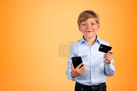 Photo for Smiling school boy with smartphone and credit card in hands, portrait on copy space yellow background. Concept of online banking and shopping - Royalty Free Image