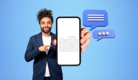 Téléchargez les photos : Arabe homme d'affaires pointent du doigt grand écran de téléphone vide, bulle de message texte sur fond bleu. Concept de messager et application mobile - en image libre de droit