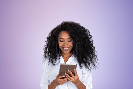 Téléchargez les photos : Portrait d'une jeune femme d'affaires afro-américaine souriante regardant un écran de tablette se tenant sur un fond violet. Concept d'internet - en image libre de droit