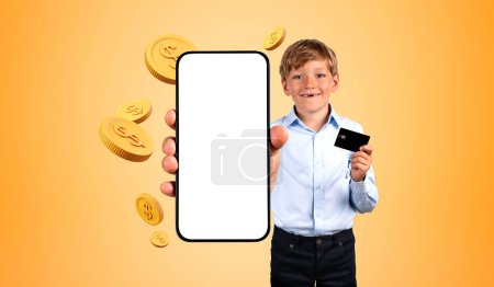 Photo for Cheerful little boy with credit card showing smartphone with mock up screen standing over orange background with dollar coins. Concept of online shopping - Royalty Free Image