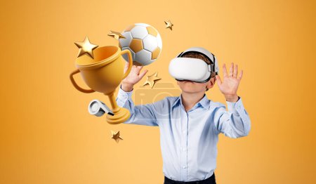 Foto de Niño en gafas VR de pie cerca de la pared naranja con trofeo y pelota de fútbol. Concepto de simulación deportiva y juego - Imagen libre de derechos