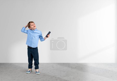 Foto de Niño inteligente soñando, mirada pensativa con el teléfono móvil en la mano en el suelo de hormigón gris, fondo blanco vacío. Concepto de carrera futura y e-learning. Copiar espacio - Imagen libre de derechos