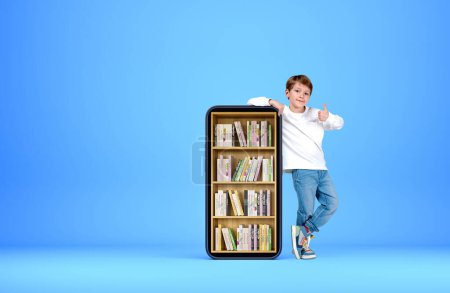 Téléchargez les photos : Enfant souriant montrant pouce debout près du smartphone avec bibliothèque numérique, livres sur rack sur fond bleu. Concept d'éducation en ligne - en image libre de droit