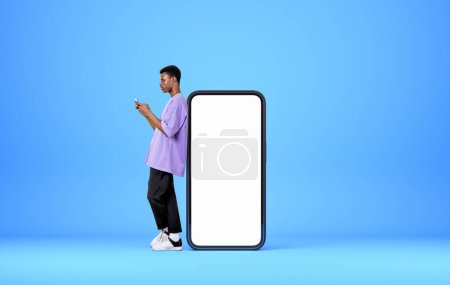 Téléchargez les photos : Homme afro-américain avec téléphone mobile, pleine longueur près de l'appareil maquette l'affichage de l'espace de copie, fond bleu clair. Concept de médias sociaux et de communication - en image libre de droit