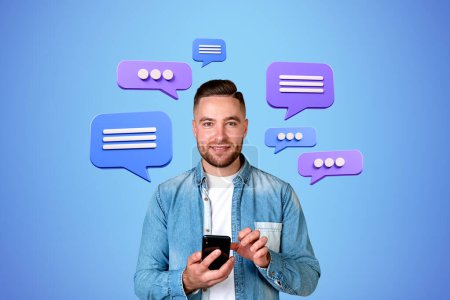 Photo for Smiling bearded young man in casual clothes using smartphone over blue background with speech bubbles. Concept of social media and chatting - Royalty Free Image
