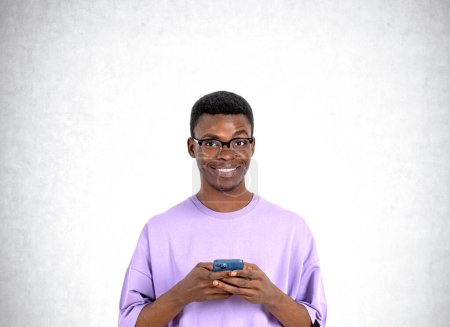 Foto de Retrato del joven afroamericano sonriente con gafas y camiseta púrpura sosteniendo teléfono inteligente cerca de la pared de hormigón. Concepto de redes sociales - Imagen libre de derechos