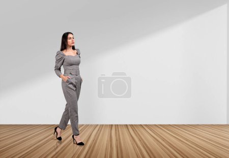 Téléchargez les photos : Femme d'affaires réfléchie marchant avec les mains dans la poche sur le plancher de bois franc, blanc espace de copie fond blanc. Concept de stratégie financière et d'investissement - en image libre de droit