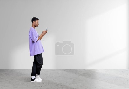 Foto de Hombre negro americano de pie con teléfono inteligente, perfil de longitud completa sobre fondo blanco, piso de hormigón gris. Concepto de redes sociales. Copiar espacio - Imagen libre de derechos