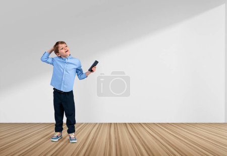 Téléchargez les photos : Enfant intelligent rêvant, look réfléchi avec smartphone à la main sur plancher de bois franc, fond blanc vide. Concept de carrière et d'éducation. Espace de copie - en image libre de droit
