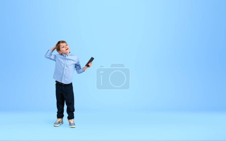 Foto de Niño pequeño soñando y mirada reflexiva sosteniendo teléfono inteligente en la mano sobre fondo azul vacío. Concepto de hobby y educación futura. Copiar espacio - Imagen libre de derechos