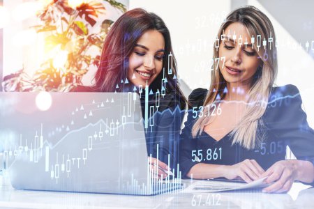 Foto de Two office woman with device, analyzing financial papers on table, double exposure with stock market hologram with lines and numbers, office room. Concept of teamwork - Imagen libre de derechos