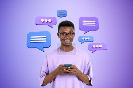 Téléchargez les photos : Joyeux jeune homme afro-américain dans des lunettes à l'aide d'un smartphone sur fond violet avec des bulles de parole. Concept de médias sociaux et de bavardage - en image libre de droit