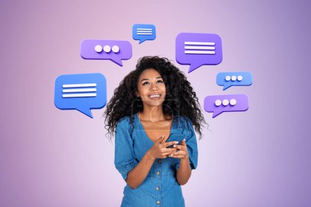 Foto de Mujer negra sonriente sosteniendo un teléfono, mirando burbujas de mensajes de texto sobre fondo púrpura. Concepto de redes sociales, comunicación y red en línea - Imagen libre de derechos