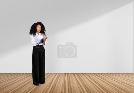 Téléchargez les photos : Une femme d'affaires afro-américaine portant des vêtements formels tient un carnet près d'un mur blanc vide en arrière-plan. Sol en bois de chêne. Concept de processus de travail, prendre des notes pour le calendrier quotidien - en image libre de droit