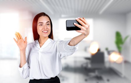 Téléchargez les photos : Heureuse femme d'affaires avec smartphone, faire appel vidéo agitant la main sur fond flou de la salle de bureau. Concept de réseau d'affaires en ligne - en image libre de droit