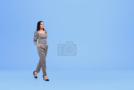 Photo for Businesswoman in jumpsuit walking hands in pocket, pensive look on blue background. Concept of career development and education. Copy space - Royalty Free Image
