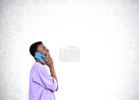 Foto de Hombre de negocios guapo afroamericano con ropa casual está hablando en el teléfono inteligente cerca de la pared de hormigón vacío en el fondo. Concepto de proceso de trabajo, trabajo a distancia, conferencia telefónica importante - Imagen libre de derechos