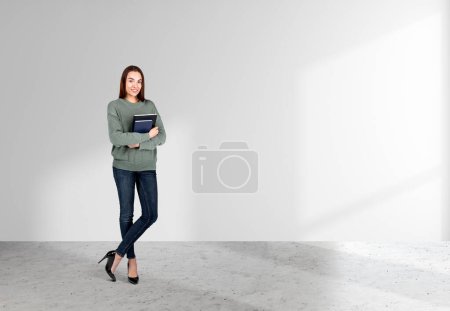 Photo for Attractive businesswoman wearing casual wear is standing holding notebook near empty white wall in background. Concrete floor. Concept of working process, taking notes for daily schedule - Royalty Free Image