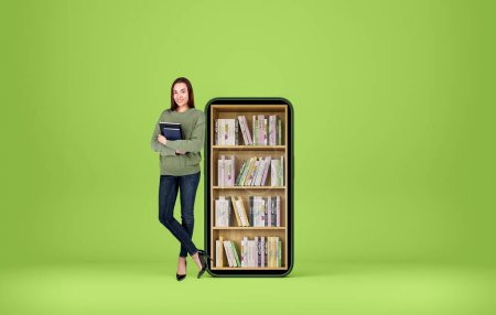 Foto de Joven mujer sonriente de pie con cuaderno en las manos, de cuerpo entero sobre fondo verde. Smartphone con libros en estante. Concepto de e-learning. Copiar espacio - Imagen libre de derechos
