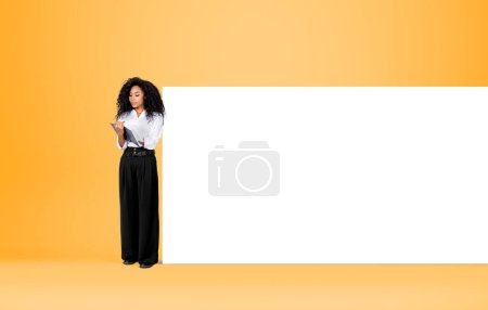 Photo for Black pensive businesswoman take note in notebook, full length standing near mockup blank whiteboard. Concept of business offer - Royalty Free Image
