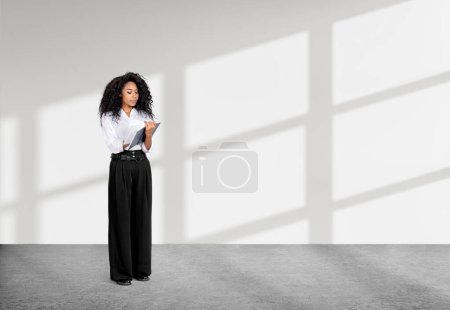 Foto de La mujer de negocios afroamericana que usa ropa formal está de pie sosteniendo un cuaderno cerca de una pared vacía con luz solar en el fondo. Piso de hormigón. Concepto de proceso de trabajo, tomando notas para el horario diario - Imagen libre de derechos
