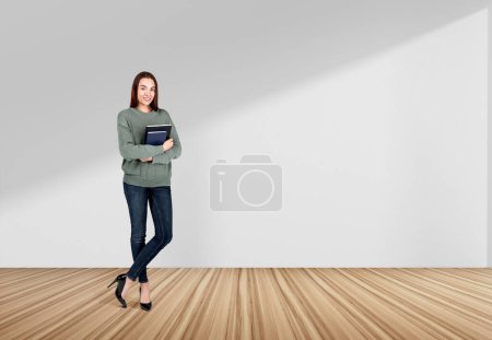 Foto de Atractiva mujer de negocios con ropa casual está de pie sosteniendo portátil cerca de la pared vacía de hormigón en el fondo. Suelo de madera de roble. Concepto de proceso de trabajo, tomando notas para el horario diario - Imagen libre de derechos